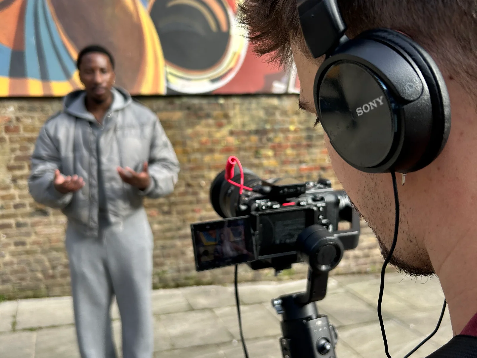 A camera operator in headphones films a contributor speaking to camera in front of a brick wall and a mural.