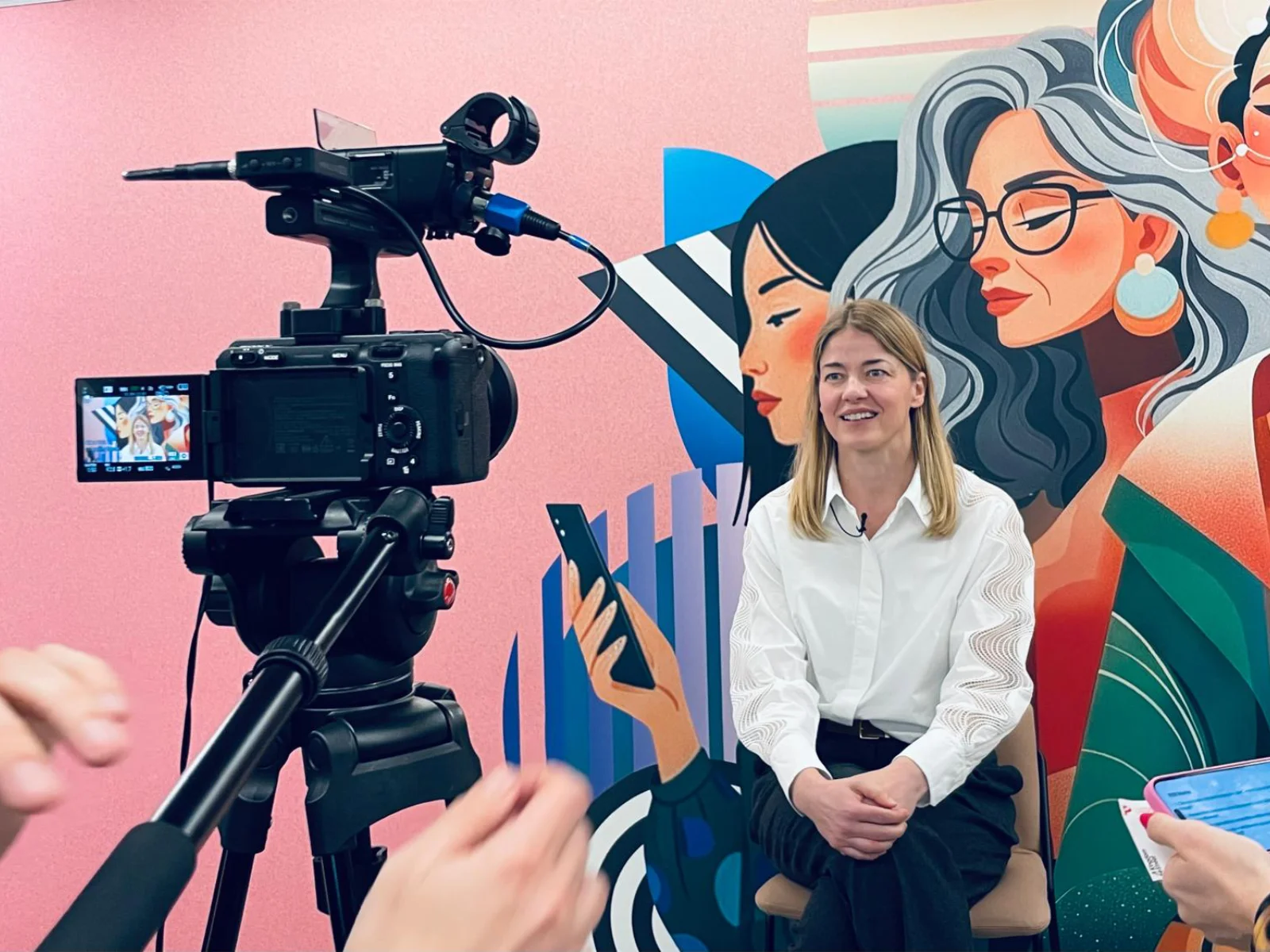 An interview in progress: a woman seated against a bright mural, framed on the screen of a tripod-mounted camera.