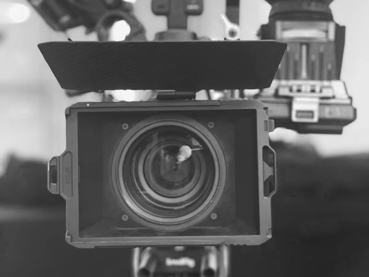 A cinema camera on a tripod seen head on in black and white, matte box and lens filling the frame.