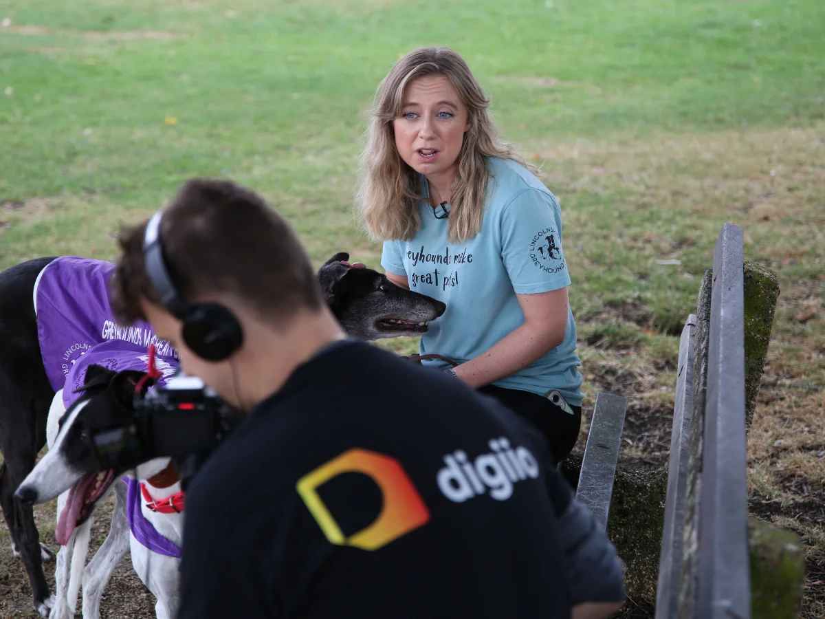A digiio crew member films an on-location interview with a charity volunteer and two greyhounds in a park.