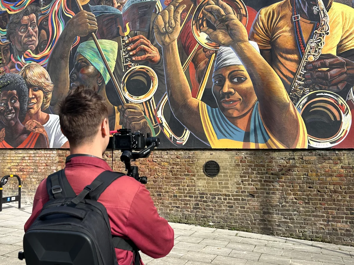 A camera operator on a gimbal films a large street mural on a brick wall in bright daylight.
