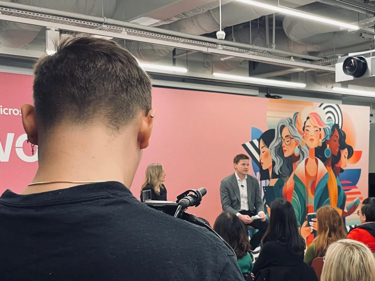 A crew member films a panel discussion on stage in front of a seated audience at a corporate event.
