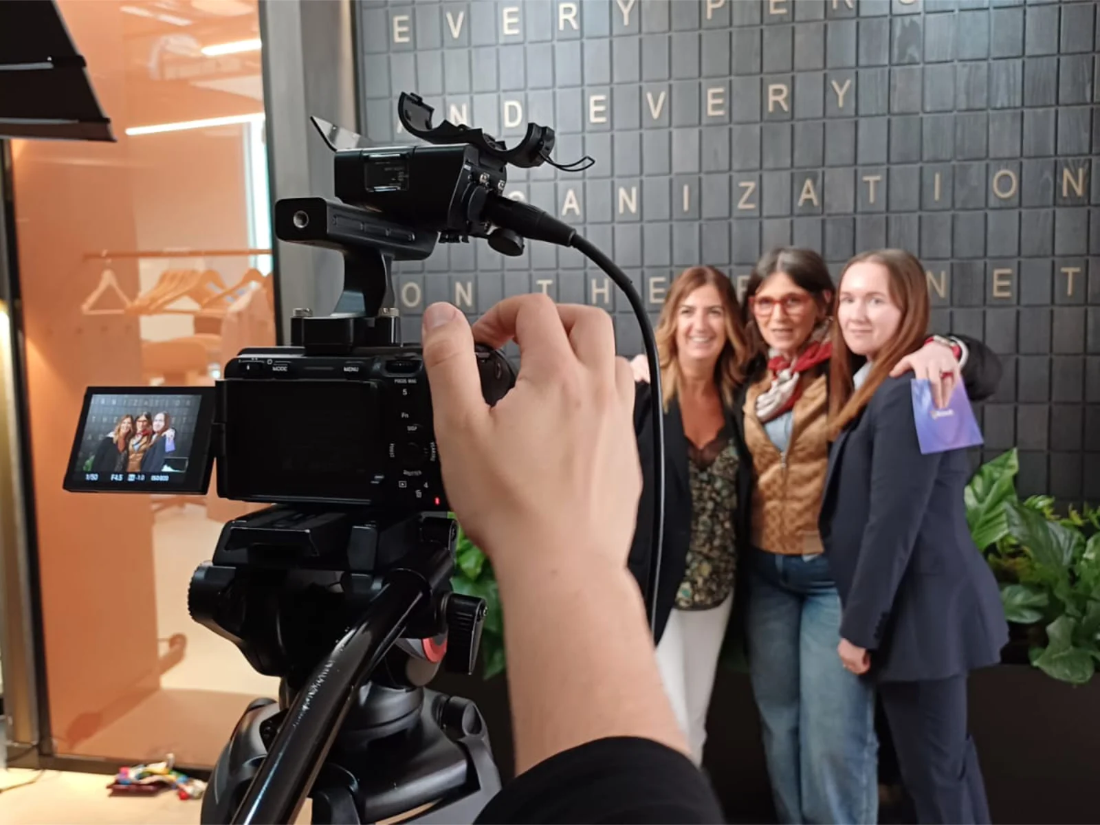 Three digiio colleagues stand together against a lettered wall while a camera operator frames the shot on a tripod-mounted camera.